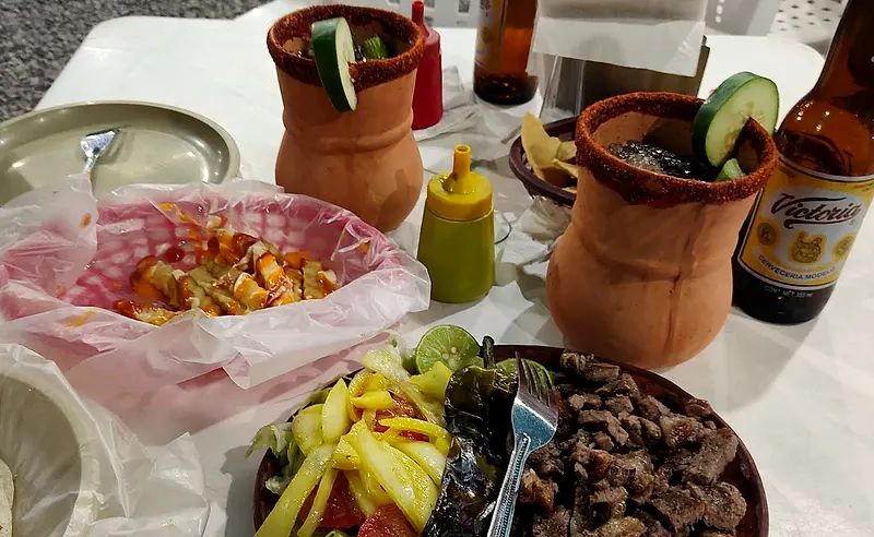 Carne asada picada con cebolla asada, chile toreado y limón, junto a papas, cantaritos y una Victoria en la mesa