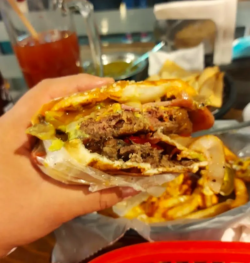 Hamburguesa doble de Lucky recién mordida: dos carnes a la parrilla con queso, tocino y aguacate, con papas y un tarro michelado atrás